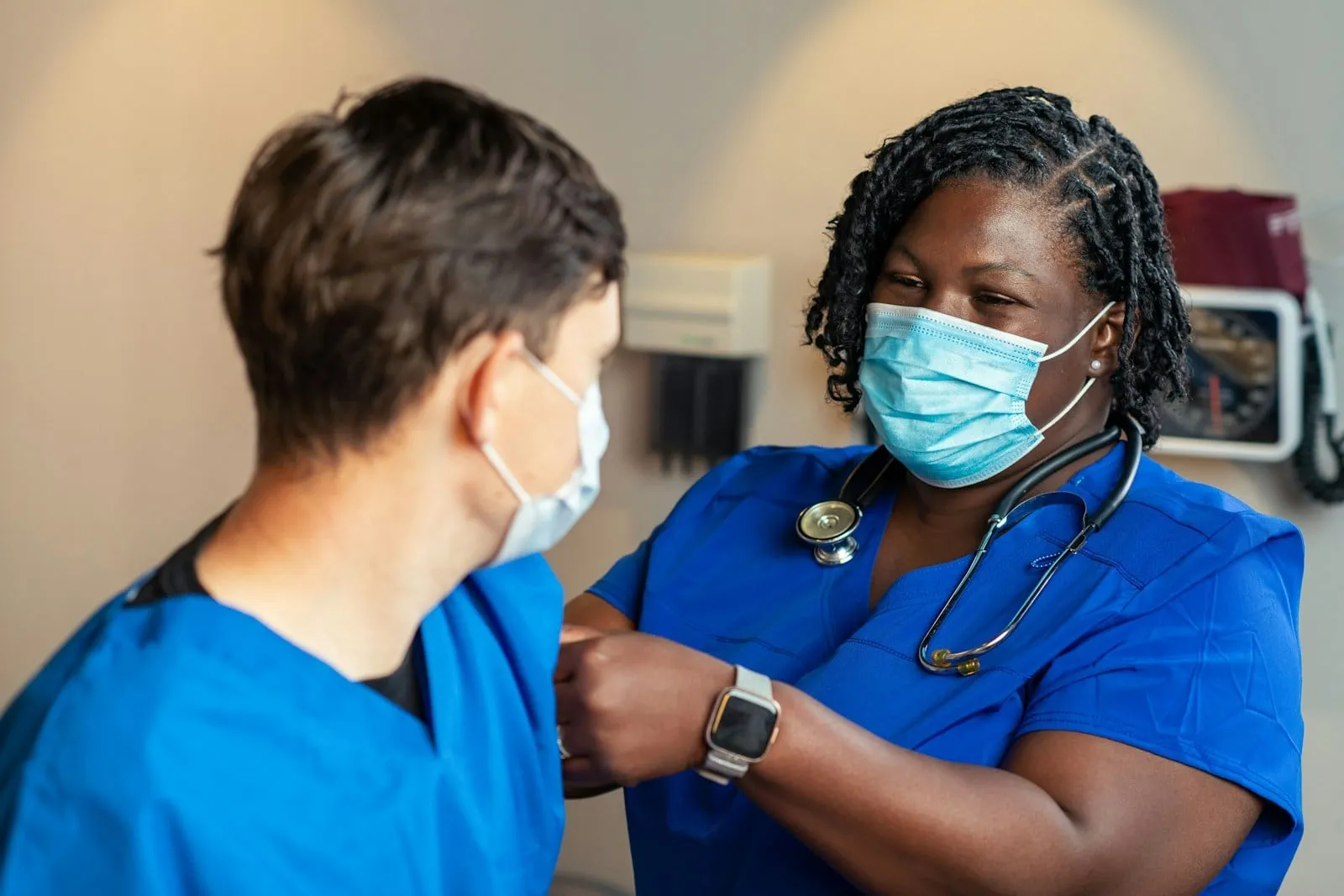 A nurse in a mask and scrubs checking a patient's arm