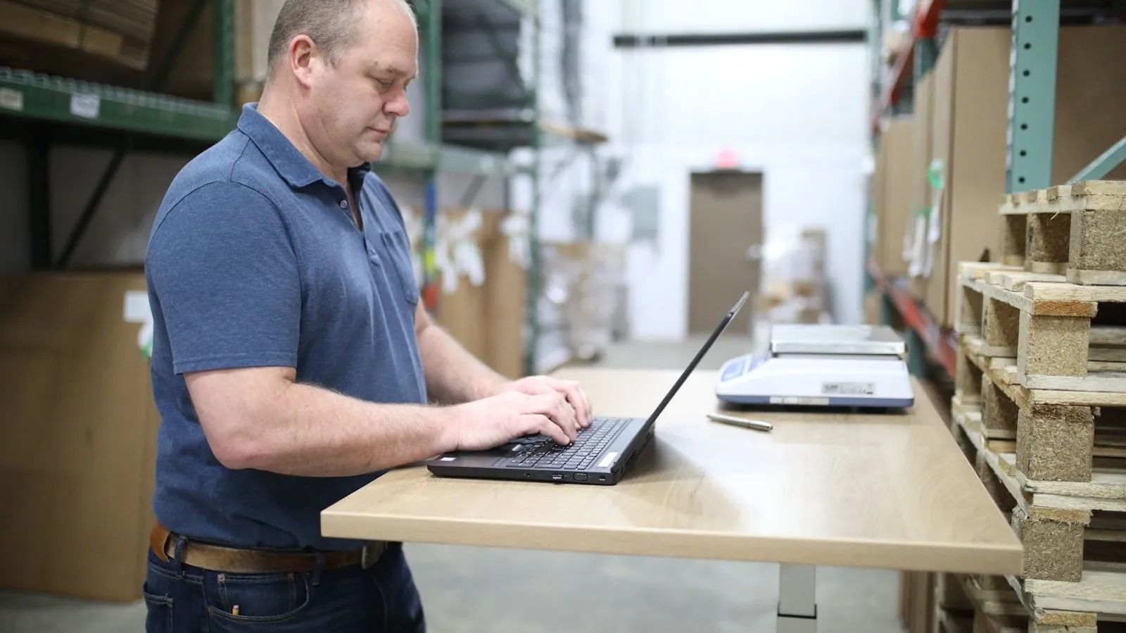A warehouse worker typing on a laptop at a standing desk beside stocked shelves