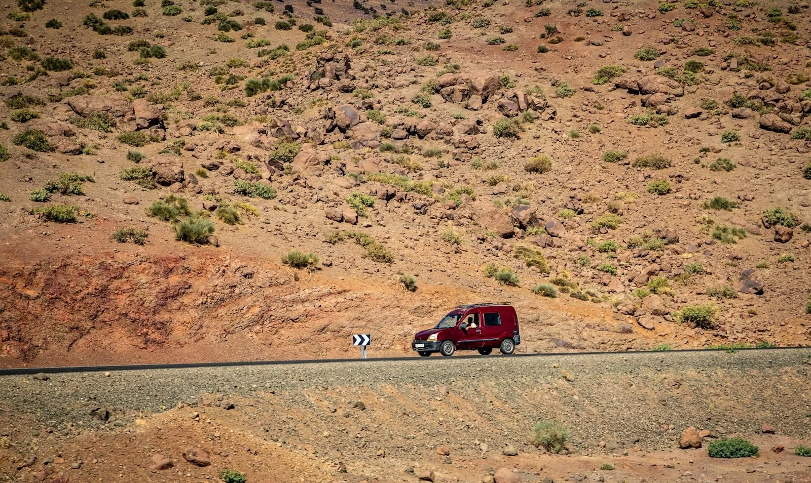 A red van driving along a remote road below a rocky hillside