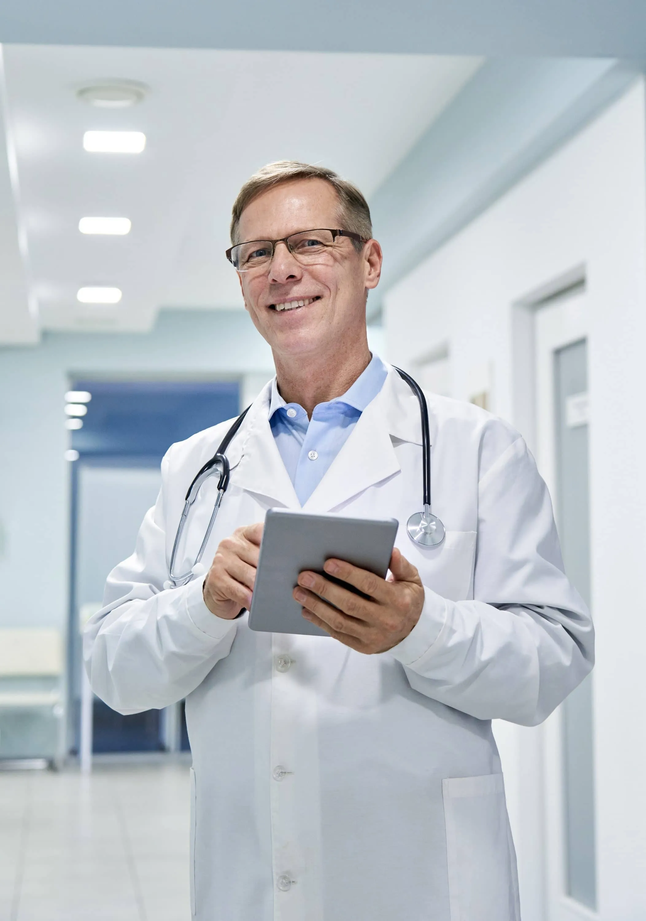 A physician in a white coat holding a tablet in a clinic hallway
